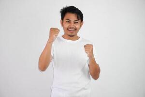 confiante alegre bonito ásia homem elevação mãos acima vestindo branco t camisa vencedora gesto aperto punhos. sente felicidade mostrar punho acima sucesso isolado em branco fundo foto