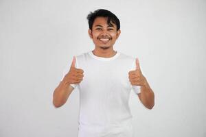 retrato do alegre ásia homem dentro vestindo branco t camisa sorridente e mostrando polegares acima às Câmera isolado sobre branco fundo foto