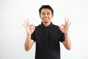 jovem ásia homem sorridente amigáveis enquanto dando Está bem dedo placa vestindo Preto pólo t camisa isolado em branco fundo foto