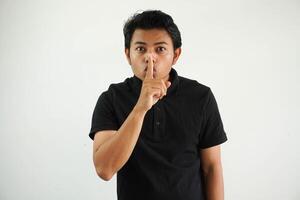 jovem ásia homem dedo toques boca guardando uma segredo ou Perguntando para silêncio vestindo Preto pólo t camisa isolado em branco fundo foto