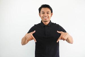 jovem ásia homem sorridente às a Câmera e apontando abaixo vestindo Preto pólo t camisa isolado em branco fundo. foto