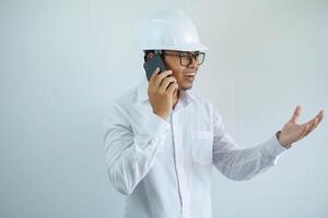 jovem ásia masculino engenheiro vestindo branco Difícil chapéu estava falando em uma célula telefone para construção trabalhos isolado em branco fundo, cópia de espaço. foto