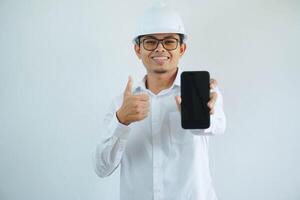 jovem ásia arquiteto homem vestindo construtor segurança capacete sobre isolado fundo fazendo feliz polegares acima gesto e segurando Móvel telefone com mão. foto