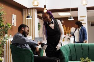 sorridente africano americano homem turista segurando crédito cartão pagando conta enquanto em repouso relaxante dentro hotel salão área. amigáveis fêmea garçonete segurando pos terminal servindo convidado às hotel lobby foto