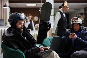 dois visitantes sentar confortavelmente usando digital dispositivos enquanto snowboard equipamento sugere perto inverno Esportes. imagem mostrando homem e mulher surfar em computador portátil e Móvel telefone dentro esqui hotel salão. foto