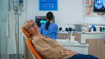 retrato do Senior mulher olhando em Câmera esperando para estomatologista dentro dental clínica. idosos paciente deitado em estomatológico cadeira sorridente às Webcam enquanto enfermeira trabalhando em computador dentro fundo. foto