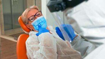 retrato em Senior paciente vestindo proteção mascarar com dentes dor durante coronavírus pandemia às clínica. assistente e médico com macacão face escudo mascarar luvas examinando mulher a partir de dental cadeira foto