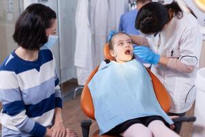 pequeno menina dentro a curso do dental tratamento a partir de jovem dentista dentro escritório. odontologia especialista durante criança cavidade consulta dentro estomatologia escritório usando moderno tecnologia. foto