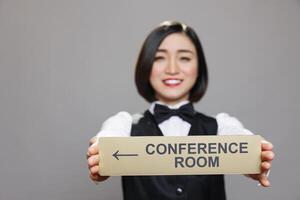 sorridente hotel administrador segurando dentro mãos inoxidável aço conferência quarto direção sinalização. mulher recepcionista mostrando escritório sala de reuniões aço placa e olhando às Câmera foto