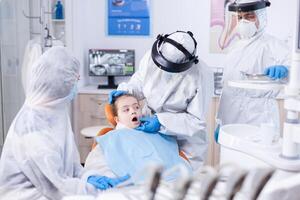 pequeno menina com boca aberto sentado em dental cadeira vestido dentro ppe roupa Como segurança precaução dentro a curso do coronavírus. dentista dentro coronavírus terno usando curvado espelho durante dentes exame do criança. foto