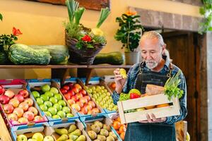Senior verdureiro trabalhando às a mercado segurando uma caixa contendo fresco frutas e legumes - Comida varejo conceito foto