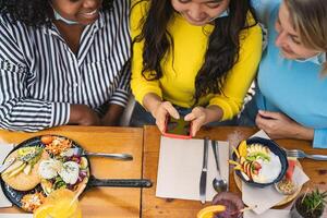 multirracial amigos assistindo em Móvel Smartphone enquanto tendo saudável almoço dentro café Escovar Barra durante corona vírus surto - Comida e tecnologia conceito foto