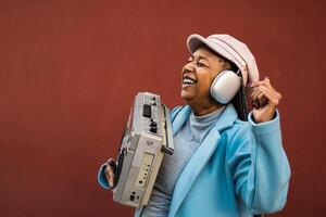 feliz na moda Senior africano mulher tendo Diversão dançando enquanto ouvindo música com fones de ouvido e vintage boombox estéreo foto