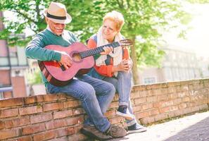 feliz Senior casal jogando uma guitarra enquanto sentado lado de fora em uma parede em uma ensolarado dia - conceito do ativo idosos tendo Diversão com guitarra - desfrutando estilo de vida durante aposentadoria foto