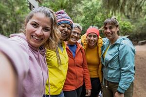 grupo do mulheres com diferente idades e etnias tendo Diversão levando selfie enquanto caminhando dentro nebuloso floresta - aventura e viagem pessoas conceito foto