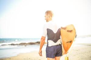 jovem surfista segurando dele prancha de surfe olhando a ondas para surfar - bonito homem em pé em a de praia às pôr do sol Treinamento para surfar - pessoas, esporte e estilo de vida conceito foto