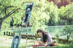 jovem artista menina desenhando esboço, projeto dentro parque perto lago - pintor mulher com tranças Penteado trabalhando em dela arte dentro a natureza ao ar livre - conceito do pessoas expressando artes foto