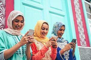 jovem muçulmano amigo usando Móvel smartphones dentro a Faculdade - feliz árabe meninas viciado para Novo tecnologia aplicativo celular para social meios de comunicação - milenar, religião, geração z e tecnologia conceito foto