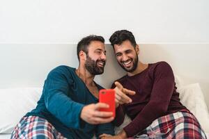 feliz gay casal tendo Diversão usando Móvel Smartphone dentro cama - homossexual amor e tecnologia conceito foto