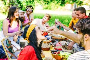 feliz amigos fazer uma piquenique dentro a jardim ao ar livre - jovem na moda pessoas tendo Diversão comendo e bebendo enquanto sentado em a Relva dentro a natureza - juventude, amizade, Comida conceito - foco em fêmea foto