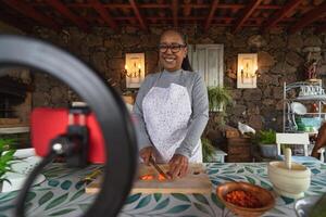 africano Senior mulher preparando Comida receita enquanto transmissão conectados com Móvel Smartphone cam para rede cozinha classe mestre canal foto