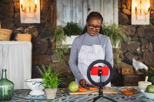 africano Senior mulher preparando Comida receita enquanto transmissão conectados com Móvel Smartphone cam para rede cozinha classe mestre canal foto