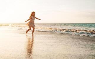 feliz criança tendo Diversão corrida em a de praia às pôr do sol - adorável pequeno menina jogando ao longo a mar água - infância e liberdade verão dias conceito foto
