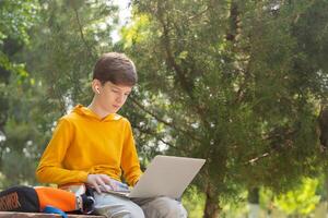 pensativo adolescente Garoto em repouso. segurando e usando uma computador portátil para trabalho em rede em uma ensolarado dia, ao ar livre. foto