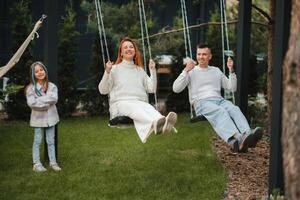 mãe e Papai estão equitação em uma balanço e lá é uma filha em pé Próximo para eles. a família é em repouso em uma balanço foto