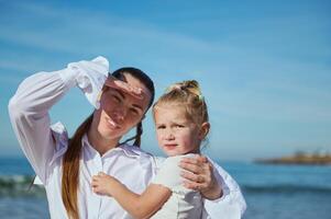 feliz carinhoso mãe segurando dela bebê menina dentro dela braços enquanto caminhando juntos em a praia, olhando para dentro a distância foto