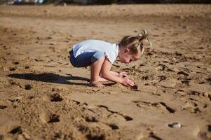 adorável bebê menina jogando com molhado areia em a praia, em pé descalço e deixando passos em a molhado arenoso de praia. foto