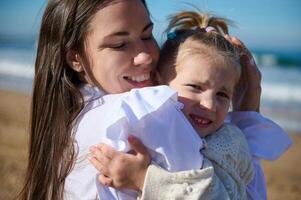 feliz mulher, jovem mãe sorridente e abraçando dela adorável pequeno criança garota, em pé juntos em arenoso de praia juntos foto