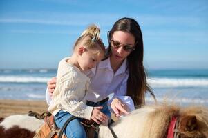 amoroso carinhoso mãe ensino dela filha para passeio uma cavalo pónei em a de praia foto