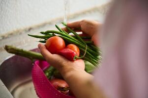 fechar-se mãos segurando fresco orgânico legumes e lavando debaixo corrida água dentro a Pia às casa cozinha foto