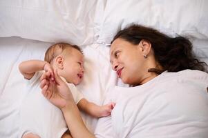 adorável pequeno bebê ter Diversão com dele mãe, sorridente para dela enquanto deitado juntos em a cama dentro a manhã. infância e maternidade sair conceito. mãe e criança juntos foto