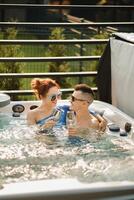 dentro verão, uma homem e uma mulher com óculos do vinho relaxar dentro a ao ar livre quente banheira foto