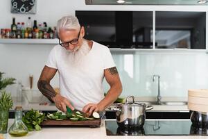 feliz Senior homem tendo Diversão cozinhando às casa - idosos pessoa preparando saúde almoço dentro moderno cozinha - aposentado estilo de vida Tempo e Comida nutrição conceito foto