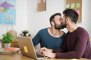 feliz gay casal se beijando enquanto usando computador portátil dentro vivo quarto às casa - lgbt amor e tecnologia conceito foto