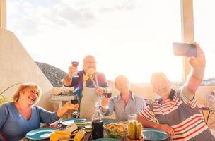 feliz idosos levando uma selfie com Móvel telefone e fazer uma churrasco em a telhado - aposentado pessoas tendo Diversão comendo e bebendo vermelho vinho rindo juntos - alegre idosos atividade conceito foto