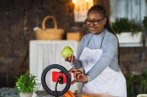 africano Senior mulher preparando Comida receita enquanto transmissão conectados com Móvel Smartphone cam para rede cozinha classe mestre canal foto