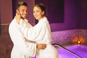 feliz jovem casal vestindo roupão de banho desfrutando a spa dia durante seus lua de mel - romântico amantes tendo relaxar dentro uma hotel spa pronto para Faz uma banheira jacuzzi - conceito sobre pessoas e bem ser foto