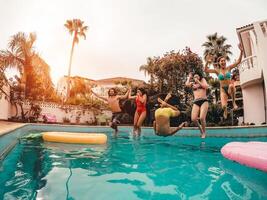 grupo do feliz amigos pulando dentro piscina às pôr do sol Tempo - louco jovem pessoas tendo Diversão fazer festa dentro exclusivo tropical casa - feriados, verão, período de férias e juventude estilo de vida conceito foto