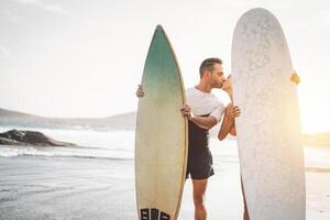 feliz casal do surfista se beijando antes para ir para surfar - desportivo pessoas tendo uma concurso momento em a de praia às pôr do sol - conceito do esporte estilo de vida, extremo, amor e relação foto