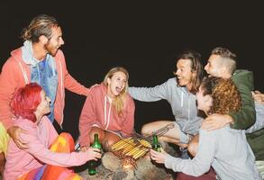 feliz amigos acampamento juntos e cozinhando milho às noite ao ar livre - jovem pessoas tendo Diversão e rindo por aí fogueira - amizade, festa, estilo de vida, selvagem conceito foto