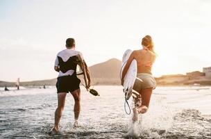 feliz surfistas corrida dentro a água às pôr do sol Tempo - jovem casal tendo Diversão surfar dentro oceano - extremo esporte e juventude cultura estilo de vida conceito foto