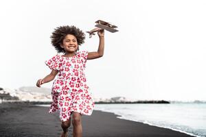 afro criança jogando com madeira brinquedo avião em a de praia - pequeno criança tendo Diversão durante verão feriados - infância e viagem período de férias conceito foto