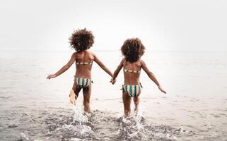 feliz irmãs corrida dentro água durante verão Tempo - afro crianças tendo Diversão jogando em a de praia - família amor e viagem período de férias estilo de vida conceito foto