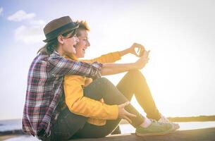 feliz lésbica casal levando uma selfie com Móvel inteligente telefone Câmera em a de praia às pôr do sol - vinheta editar - homossexualidade, diversidade, férias, viagem, lgbt, tecnologia conceito foto