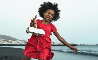 afro criança jogando com madeira brinquedo avião em a de praia - pequeno criança tendo Diversão durante verão feriados foto
