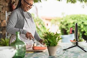 feliz afro Senior mulher preparando Comida receita enquanto vlogging conectados com Móvel Smartphone cam para rede cozinha classe mestre canal foto